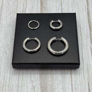 K&ouml;nnte beinhalten: Vier silberne Creolen in verschiedenen Gr&ouml;&szlig;en, die auf einem schwarzen Hintergrund pr&auml;sentiert werden. Die Ohrringe sind alle aus Metall und haben eine glatte Oberfl&auml;che.