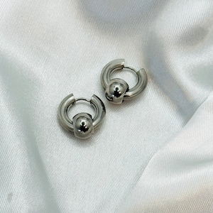 K&ouml;nnte beinhalten: Ein Paar silberne Creolen mit einem Kugeldetail. Die Ohrringe sind aus Metall und haben eine polierte Oberfl&auml;che.