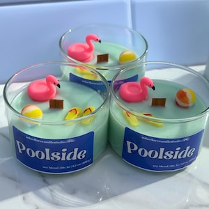 May include: Three clear glass candles with a light green wax, each decorated with a pink flamingo, a small wooden block, a beach ball, and yellow flip-flops. The candles have a blue label that reads "Poolside".