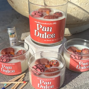 Puede incluir: Cuatro velas de vidrio con cera blanca y productos horneados decorativos en la parte superior. Las velas tienen etiquetas rojas con el texto "Pan Dulce". También hay una pequeña botella de cerillas y algunas cerillas sueltas.