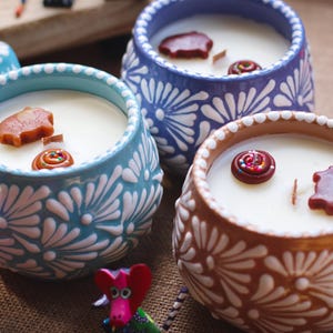 May include: Three ceramic mugs filled with white wax candles, each featuring a handle and a white floral pattern on a light blue, dark blue, or brown background. The candles contain small decorative wax shapes including pigs and swirls.
