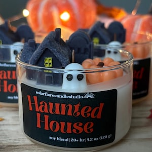 May include: A white soy candle with a black label that reads "Haunted House" and "soy blend | 20+ hr | 4.2 oz (120 g)". The candle is decorated with a small white ghost, a small orange pumpkin, and a small black house.