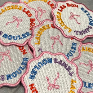 May include: A collection of circular embroidered patches with scalloped pink edges. Each white patch features a pink ribbon graphic in the center and the text LAISSEZ LES BONS TEMPS ROULER embroidered in alternating yellow, red, blue, and purple thread.
