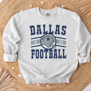 Puede incluir: Una sudadera gris con un gráfico de estilo vintage de un casco de fútbol y el texto "Dallas Football" con los números "19" y "60" a cada lado del casco.