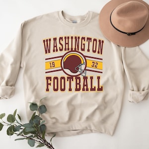 May include: A beige sweatshirt with the text "WASHINGTON FOOTBALL" in maroon and gold lettering. The number "19" and "32" are on either side of the text. A maroon and gold football helmet is above the text.