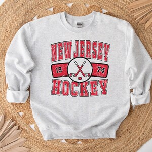 May include: A gray sweatshirt with a red and white graphic print that says "New Jersey Hockey" with crossed hockey sticks and the numbers 19 and 74.