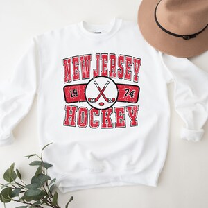 May include: White sweatshirt with a red and black graphic that says "New Jersey Hockey" with crossed hockey sticks and the numbers 19 and 74.