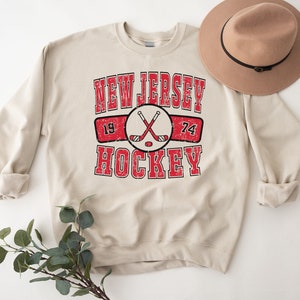 May include: A beige sweatshirt with a vintage-style graphic design that says "New Jersey Hockey" with crossed hockey sticks and the numbers 19 and 74.