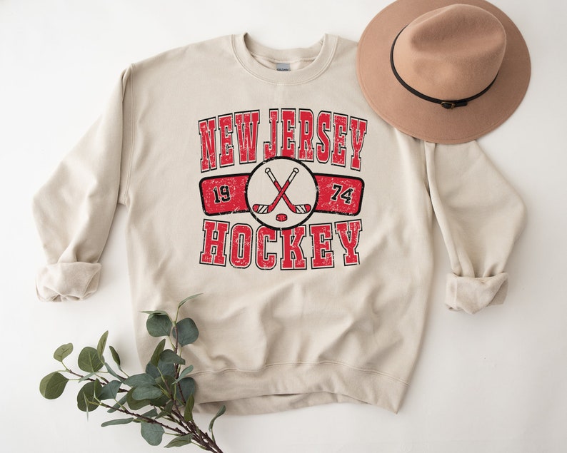 May include: A beige sweatshirt with a vintage-style graphic design that says "New Jersey Hockey" with crossed hockey sticks and the numbers 19 and 74.