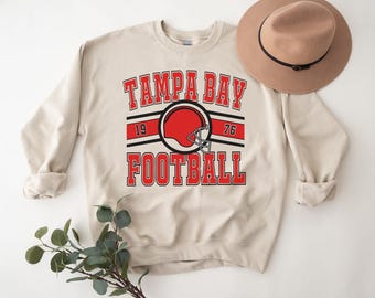 Pode incluir: Uma camisa de moletom bege com uma estampa gráfica vermelha, preta e branca que diz "Tampa Bay Football" com um gráfico de capacete de futebol vermelho.