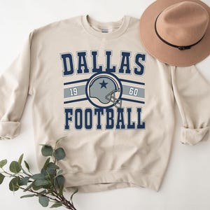 Puede incluir: Una sudadera beige con un gráfico de Dallas Football azul y blanco. El gráfico incluye un casco de fútbol con una estrella y los números 19 y 60.