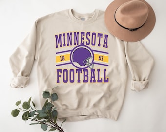 May include: A beige sweatshirt with a purple and yellow graphic that says "Minnesota Football" with a purple football helmet graphic and the numbers "19" and "61" on the graphic.