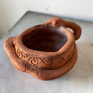 Op de afbeelding: Een kleine, met de hand gevormde terracotta kleikom met een kronkelend slangontwerp rond de buitenrand. Het oppervlak heeft een getextureerde, aardebruine afwerking met ingesneden spiraalpatronen en een handvat in de vorm van een slangenkop.