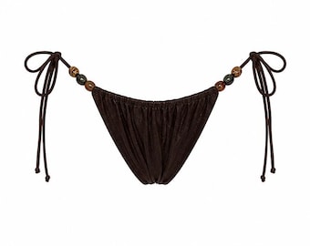 May include: A dark brown bikini bottom featuring a gathered fabric design and side ties. Each side tie is accented with two decorative beads, one dark brown and one metallic, providing a subtle contrast to the solid dark brown fabric.