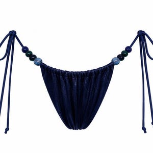 May include: A dark blue bikini bottom with a shimmering, metallic fabric finish. The sides feature thin, adjustable tie strings adorned with a series of three round beads in shades of blue, green, and dark teal on each side of the garment.