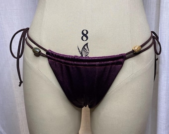 May include: A dark purple bikini bottom featuring a gathered front panel and thin side ties. The side strings are accented with small, smooth, oval-shaped beads in earthy tones. The garment is displayed on a beige mannequin torso.