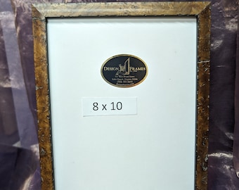 May include: A picture frame, measuring 20.3 x 25.4 cm, with a distressed gold finish. The frame has a white mat and a label that reads "DESIGN FRAMES". The frame is displayed against a purple fabric background.