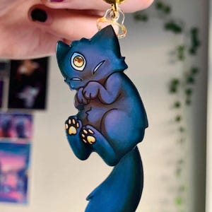 May include: A blue cat keychain with a gold-colored ring. The cat has a worried expression and yellow eyes. The cat's paws are yellow. The keychain is hanging in front of a blurred background.