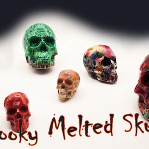 May include: A collection of colorful, melted skull sculptures in various sizes and hues, including green, red, and multicolored. The phrase "Spooky Melted Skulls" is written in brown script below the skulls.