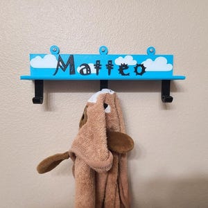 May include: A blue wall-mounted shelf with the name "Matteo" in black script, decorated with white clouds. Two black hooks hang below, supporting a brown hooded towel shaped like a dog.
