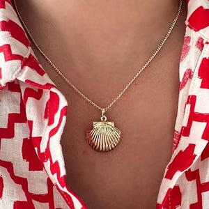 May include: A gold chain necklace with a seashell pendant. The necklace is worn by a person wearing a red and white patterned shirt.