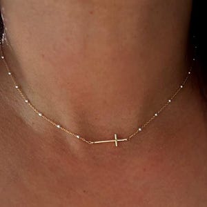 Puede incluir: Un delicado collar de oro con un pequeño colgante de cruz lateral. El collar tiene una fina cadena con pequeñas cuentas blancas espaciadas uniformemente. La cruz es simple y elegante, adecuada para el uso diario.