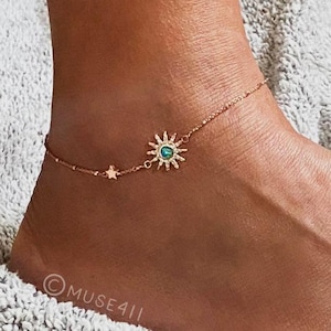 May include: A gold-colored anklet featuring a sunburst charm with a central turquoise-colored stone and a small star charm. The delicate chain is worn around the ankle. The anklet is displayed on a light gray, textured surface.