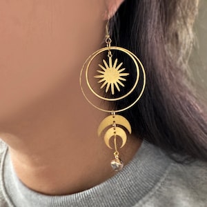 Può includere: Orecchini dorati con un designo a forma di sole e luna. Il sole è una stella ritagliata con un cerchio intorno. La luna è a forma di mezzaluna con un ciondolo di cristallo.