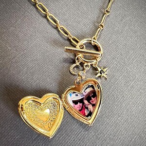 May include: A gold heart-shaped locket necklace with a toggle clasp. The locket opens to reveal a photo of two people. The necklace also features a small star charm and a crescent moon charm.