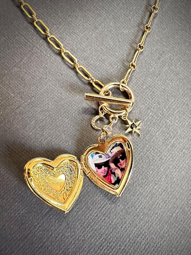 May include: A gold heart-shaped locket necklace with a toggle clasp. The locket opens to reveal a photo of two people. The necklace also features a small star charm and a crescent moon charm.