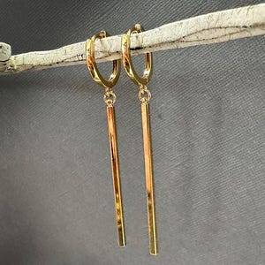 Könnte beinhalten: Ein Paar goldfarbene Creolen mit langen, rechteckigen Stäben als Anhänger. Die Ohrringe hängen an einem hellen, strukturierten Ast vor einem grauen Hintergrund. Die Reifen und Stäbe haben eine glänzende, reflektierende Oberfläche.