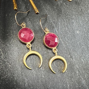 May include: Gold-coloured earrings featuring a round, faceted, magenta gemstone and a crescent moon charm. The earrings hang from a gold hook. The background is a dark, textured surface.