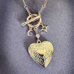 May include: A gold necklace with a heart-shaped locket, a crescent moon charm, and a star charm with a black stone. The necklace has a toggle clasp.