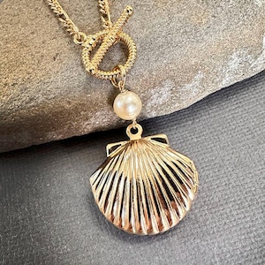 Puede incluir: Un collar de cadena dorada con cierre de palanca y un colgante de medallón de concha. El medallón está decorado con una perla.