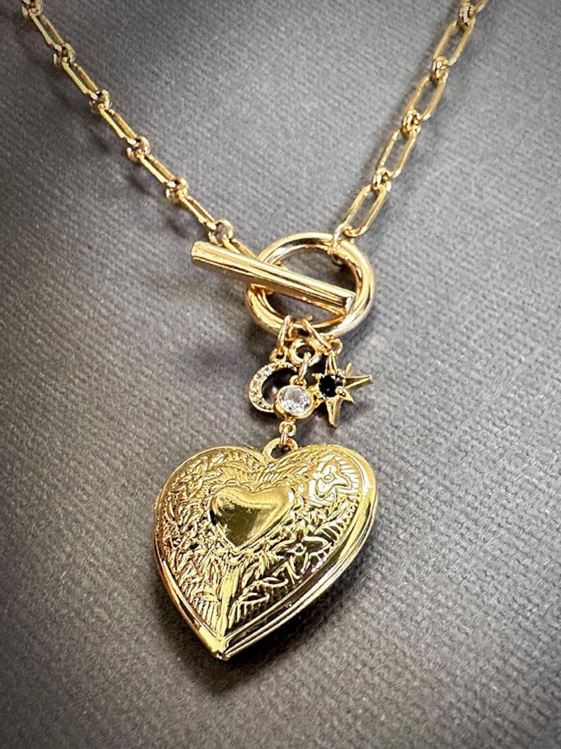 May include: A gold chain necklace with a heart-shaped locket pendant. The locket has a detailed floral design and a small charm with a star and a crystal.