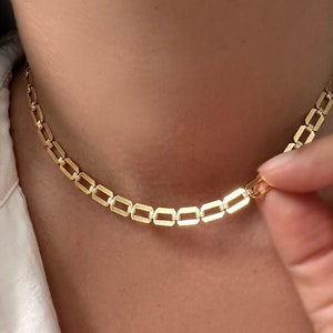 May include: A gold chain necklace with a square link design. The necklace is worn by a person, but the person's face is not visible.