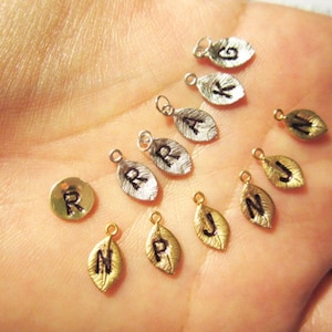 May include: Assortment of gold and silver leaf charms with different letters engraved on them. The charms are on a hand.