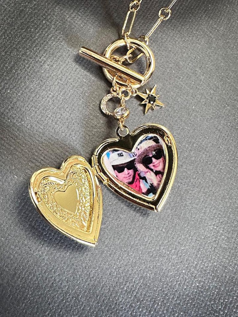 May include: Gold heart-shaped locket necklace with a photo of two people inside. The necklace has a toggle clasp and a star charm with a black stone.