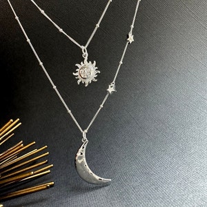 以下が含まれることがあります： 三日月型のペンダント、太陽のペンダント、星のチャーム2つが付いたシルバーネックレス。ネックレスは重ね付けになっており、繊細なチェーンを使用しています。