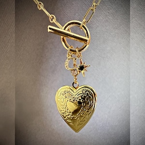May include: A gold necklace with a heart-shaped locket, a star charm, and a toggle clasp. The locket has an intricate floral design.