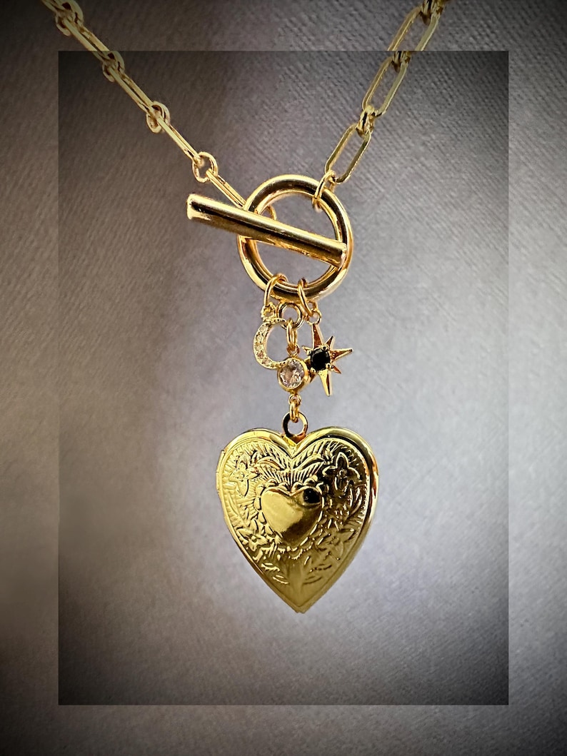 May include: A gold necklace with a heart-shaped locket, a star charm, and a toggle clasp. The locket has an intricate floral design.