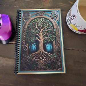 May include: Spiral-bound notebook featuring a detailed illustration of a tree with intricate roots and branches, set against a backdrop of a forest. The cover has a teal border and gold accents. A purple computer mouse is visible.