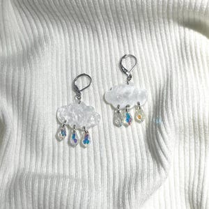 Puede incluir: Un par de pendientes en forma de nube blanca con gotas de lluvia de cristal iridiscente que cuelgan de ellos. Los pendientes están hechos de resina y tienen un gancho de metal plateado.