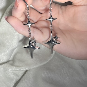 May include: A pair of silver dangle earrings with a starburst design. The earrings are made of metal and have a shiny finish.