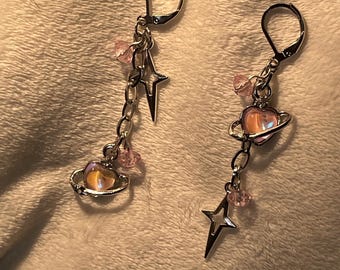 May include: A pair of silver earrings with a pink heart charm, a silver star charm, and pink crystal beads. The earrings are on a white fuzzy surface. "Pink Heart" is written below the earrings.