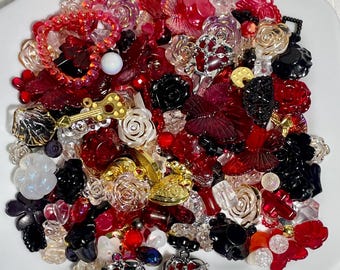 Puede incluir: Un plato blanco lleno de una variedad de cuentas, abalorios y botones de color rojo, negro y dorado. Las cuentas tienen forma de flores, corazones y otros diseños. También hay algunas cuentas de vidrio transparente y algunos abalorios plateados.