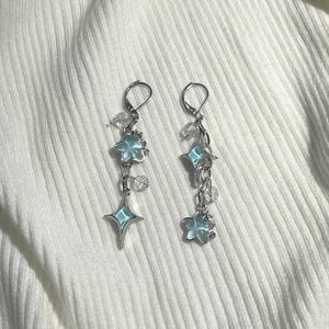 Puede incluir: Un par de pendientes de aro de plata con colgantes de estrella y flor azules. Los colgantes son de vidrio y tienen un acabado esmerilado.