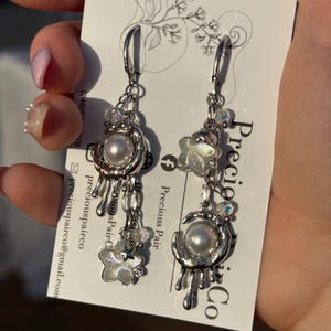 May include: A pair of silver-toned dangle earrings featuring a crescent moon charm with a central white pearl and dripping metal accents, paired with a small iridescent star charm. The earrings are displayed on a white card labeled Precious Pair Co.