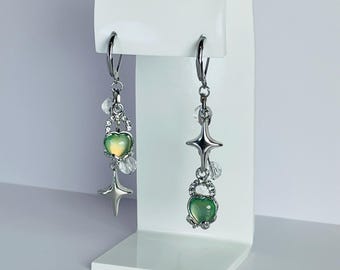 May include: A pair of silver-toned dangle earrings. Each earring features a green heart-shaped charm, a star, and clear crystal accents. The earrings are displayed on a white stand against a white background.