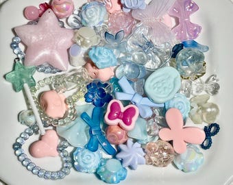 May include: A collection of pastel-colored plastic buttons and beads in various shapes, including stars, butterflies, flowers, and bows. The buttons and beads are arranged on a white plate.